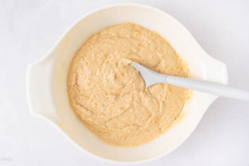 A white mixing bowl filled with thick, light brown batter. A gray spatula rests inside the bowl, partially immersed in the mixture, on a white surface.