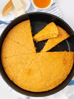 A round skillet of sliced cornbread with one piece being lifted, next to a dish of honey and a plate with butter on a striped towel.