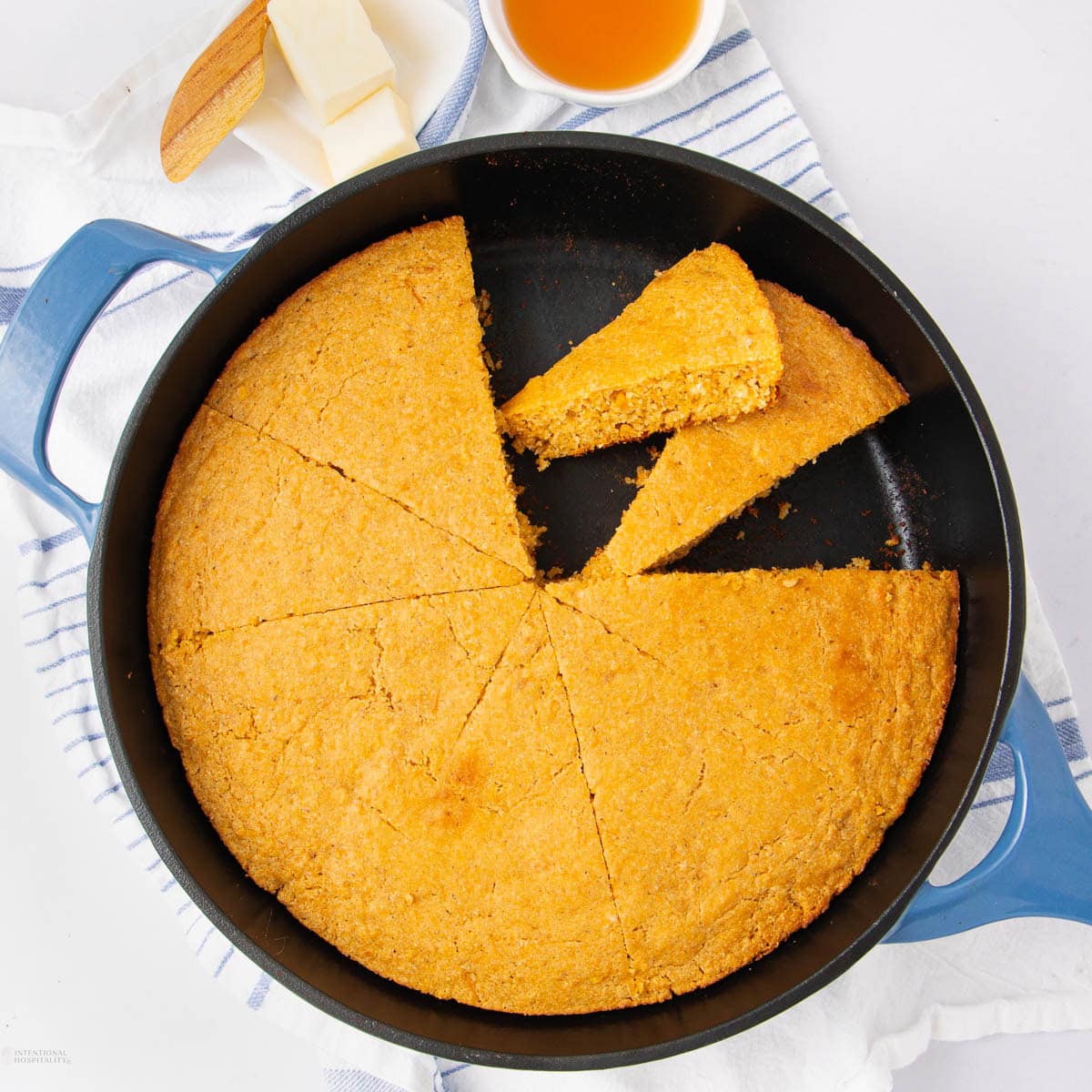 A round skillet of sliced cornbread with one piece being lifted, next to a dish of honey and a plate
