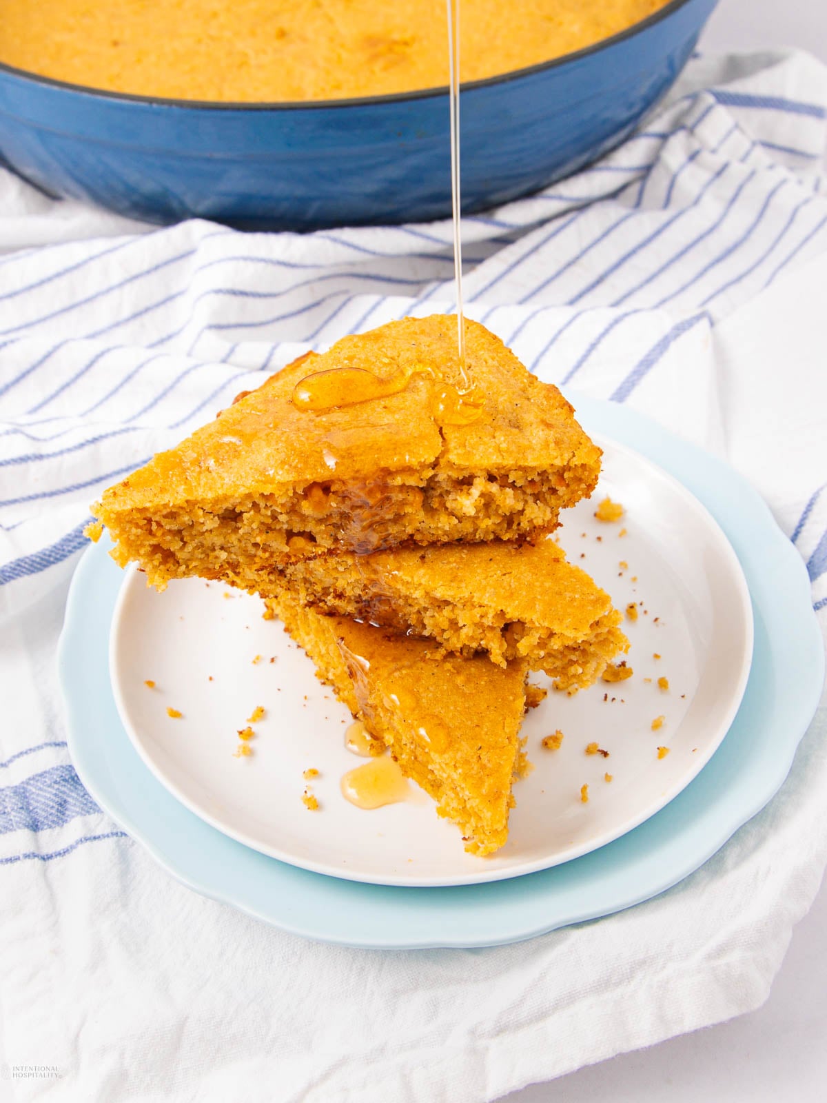 Two triangular slices of cornbread stacked on a white plate with honey being drizzled on top. The plate sits on a blue and white striped cloth with more cornbread visible in the background.