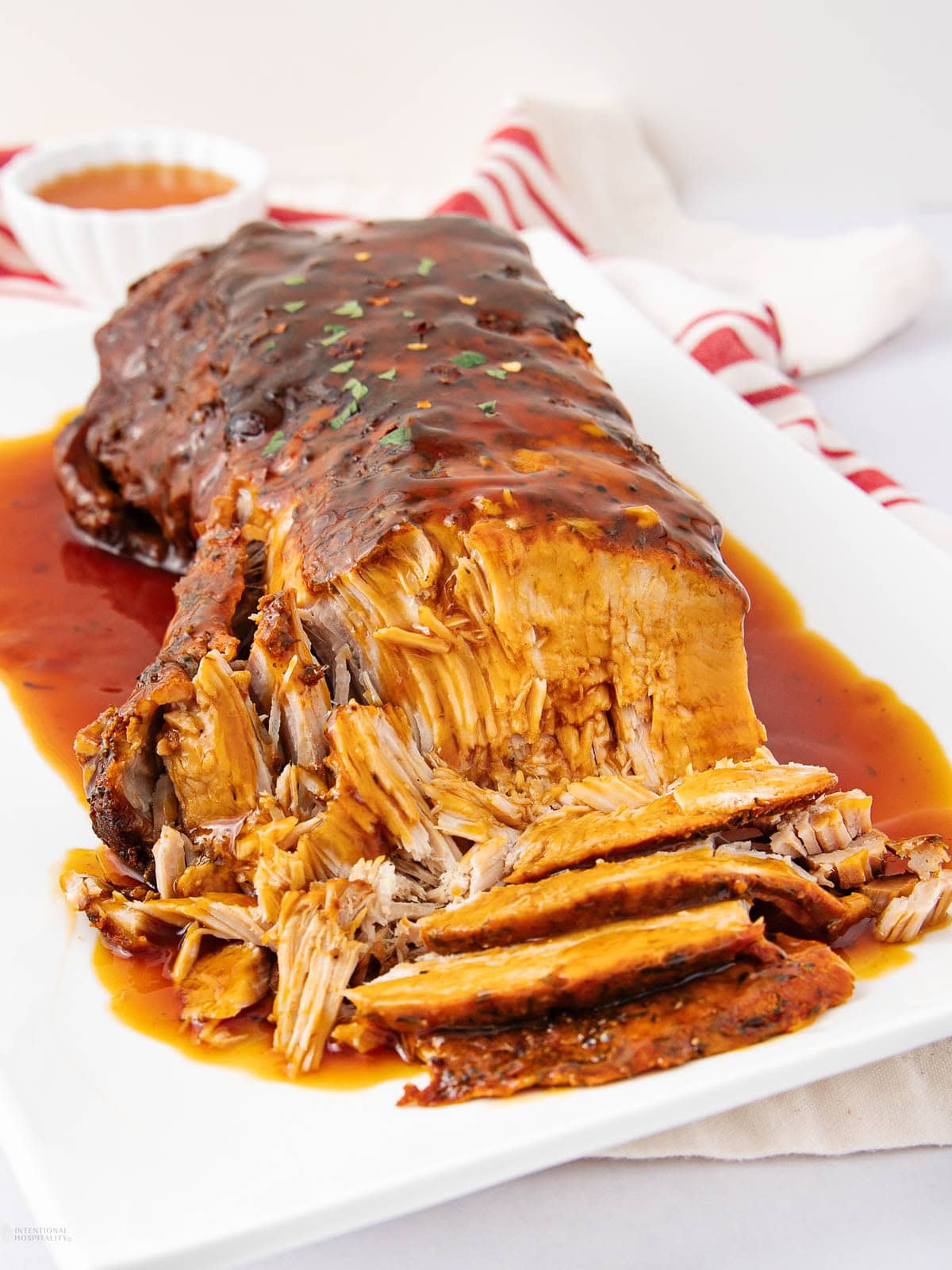 A sliced, tender pot roast topped with rich brown gravy is served on a white platter with extra sauce, accompanied by a red-and-white striped towel in the background.