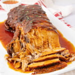 A tender, sliced pork roast covered in rich brown gravy sits on a white platter, with extra sauce pooling around the meat. A small bowl of sauce and a red-striped towel are visible in the background.