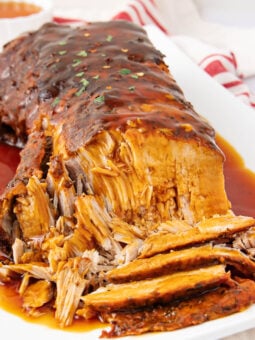 A tender, sliced pork roast covered in rich brown gravy sits on a white platter, with extra sauce pooling around the meat. A small bowl of sauce and a red-striped towel are visible in the background.
