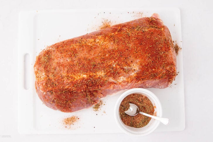Raw pork loin coated with a spice and herb rub sits on a white cutting board beside a small white bowl filled with extra seasoning mix and a spoon.