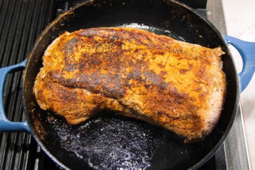 A large, seasoned beef roast is searing in a black cast iron skillet on a stovetop, with a browned and crispy crust forming on the surface.