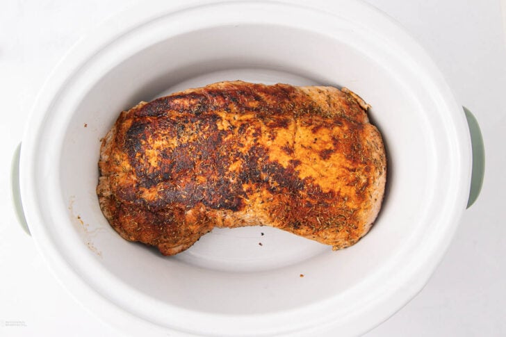 A cooked, seasoned pork roast rests inside a white oval slow cooker, viewed from above. The roast has a golden-brown, spiced crust and the slow cooker sits on a white surface.