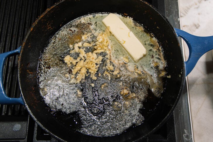 A stick of butter and chopped garlic melting in a black cast iron skillet on a stovetop.