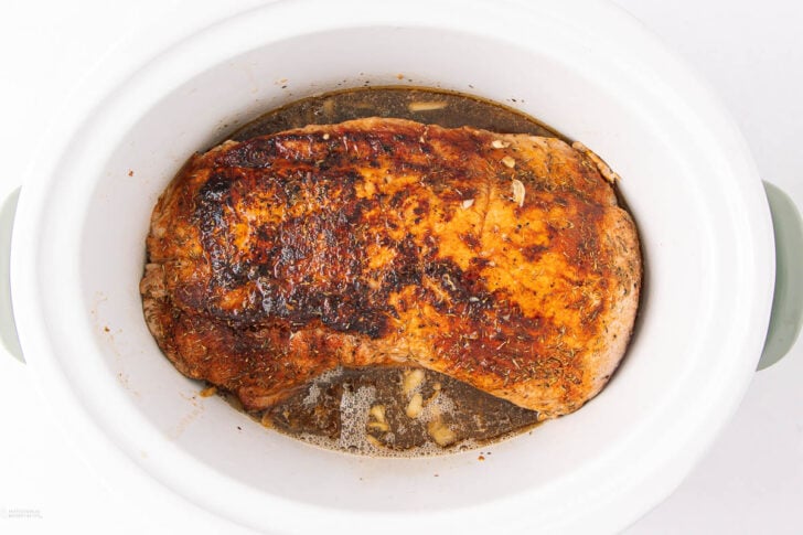 A cooked, seasoned pork roast sits in a white slow cooker with juices and herbs, showing a browned, crispy top.