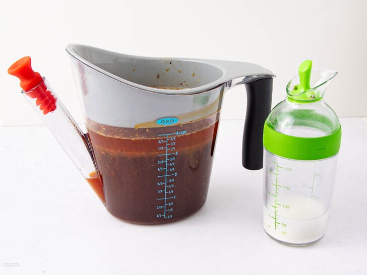 A fat separator with brown liquid and measurement markings sits next to a green-lidded bottle containing a small amount of white liquid, both on a white surface.
