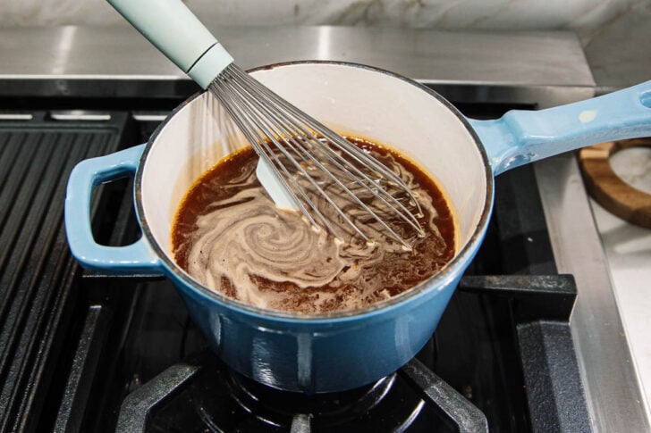 A blue pot with a brown sauce being whisked on a stovetop burner, with a metal whisk creating swirls in the liquid.
