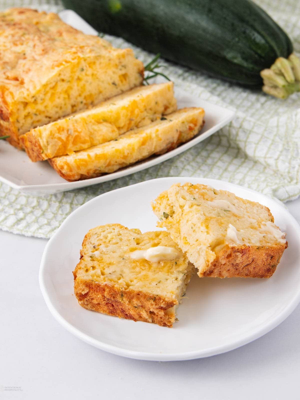 Sliced zucchini cheddar bread on a white plate, with one slice cut in half and topped with butter. More bread and a whole zucchini are visible in the background.