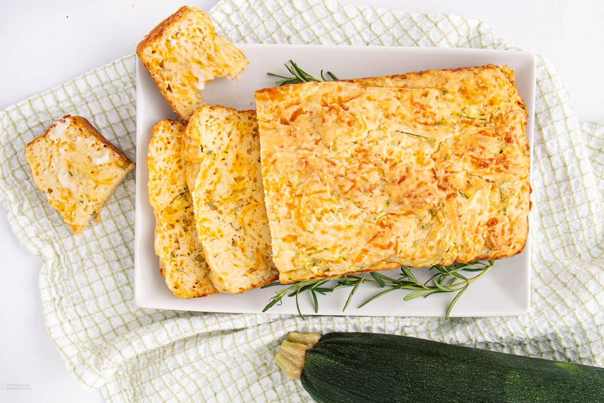 A rectangular loaf of zucchini bread, partially sliced, sits on a white plate with fresh rosemary, placed on a textured white cloth. A whole zucchini rests beside the plate.