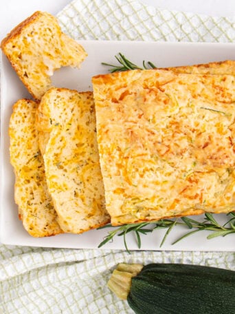 A rectangular loaf of zucchini bread, partially sliced, sits on a white plate with sprigs of rosemary. The plate is on a checkered cloth, and a whole zucchini is nearby.