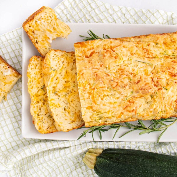 A rectangular loaf of zucchini bread, partially sliced, sits on a white plate with sprigs of rosemary. The plate is on a checkered cloth, and a whole zucchini is nearby.