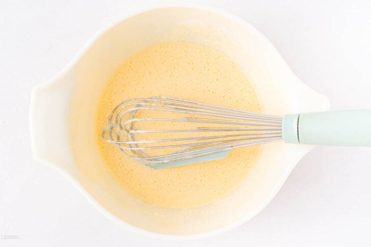 A white mixing bowl containing a light yellow batter, with a metal whisk with a pale blue handle resting inside.