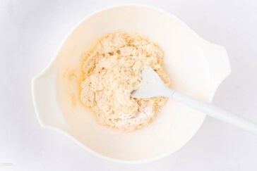 A white mixing bowl with partially mixed dough and a white spatula on a light surface.