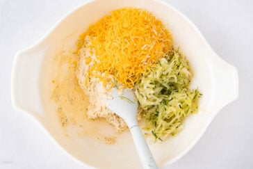 A white mixing bowl with shredded cheddar cheese, grated zucchini, and a thick batter being stirred with a white spatula.