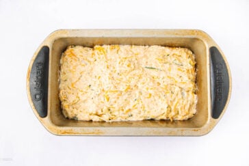 A metal loaf pan filled with unbaked cheesy bread dough, topped with shredded cheese and herbs, on a white background.