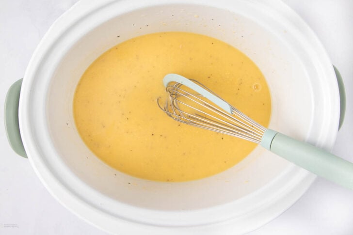 A metal whisk with a pale green handle rests in a white slow cooker filled with a creamy yellow liquid mixture.