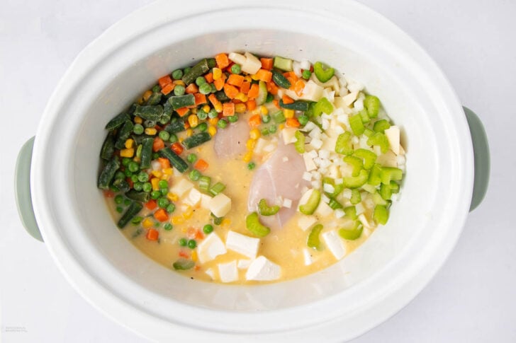 A white slow cooker containing raw chicken breasts, mixed frozen vegetables, chopped celery, diced potatoes, and cubed butter in a creamy liquid. The ingredients are uncooked and evenly distributed.