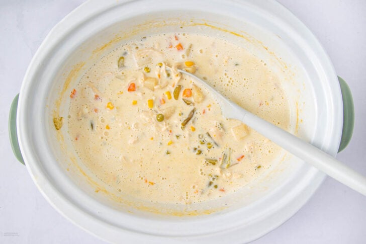 A creamy soup with mixed vegetables and shredded chicken in a white slow cooker, stirred with a white spoon.