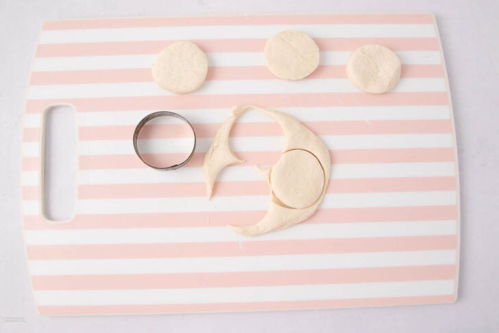 A pink and white striped cutting board with three round dough circles, a metal biscuit cutter, and a sheet of dough with a circle cut out.