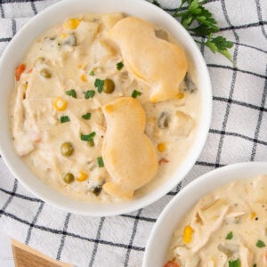 A bowl of creamy vegetable and chicken stew topped with two golden biscuit pieces, garnished with chopped green herbs, sits on a white and black checkered cloth next to another partially visible bowl.