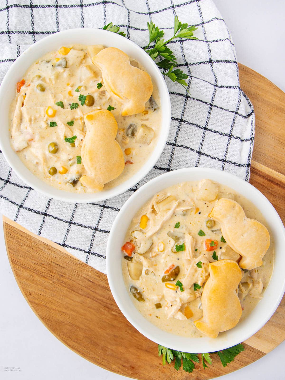 Two white bowls of creamy chicken pot pie soup with vegetables, topped with golden biscuit shapes, sit on a wooden board and a white towel with black stripes. Parsley garnish is around the bowls.