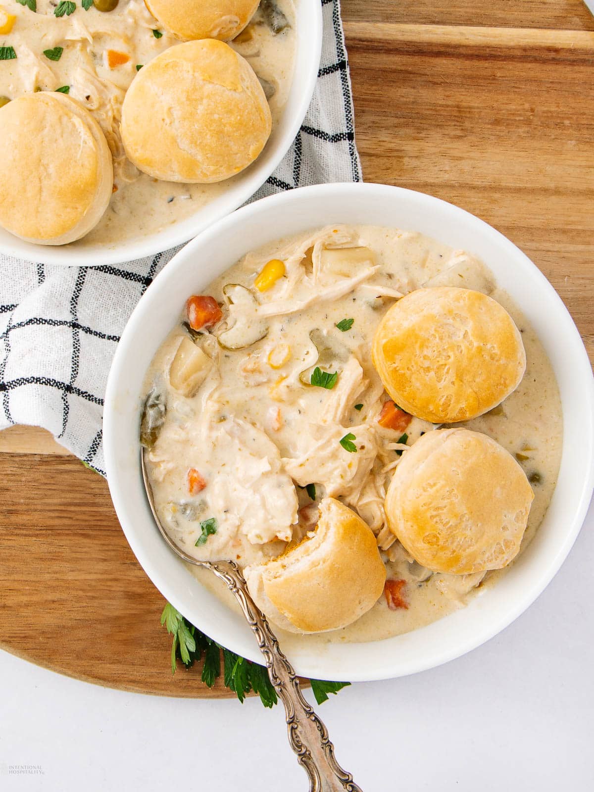 A bowl of creamy chicken and vegetable stew topped with three golden biscuits, with a spoon inside the bowl. Another bowl with biscuits is partially visible on the side, all set on a wooden surface with a striped cloth.