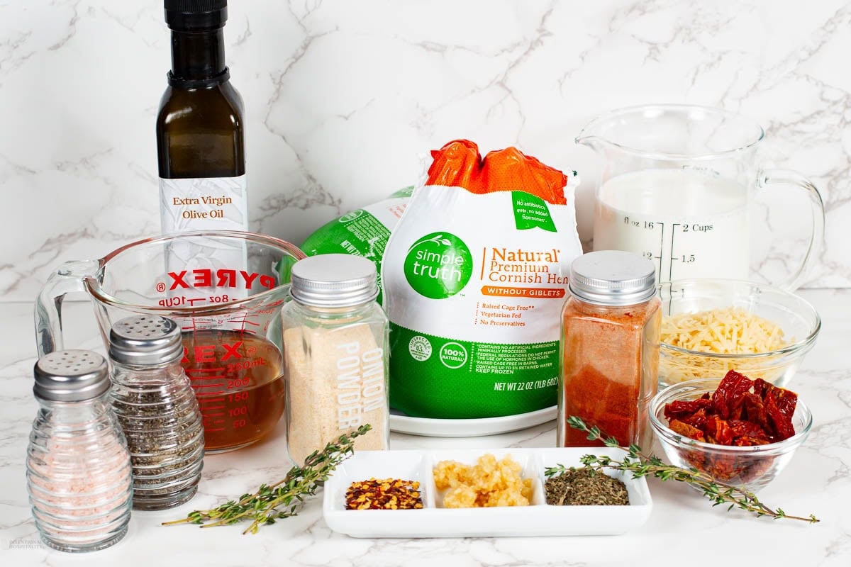 A variety of ingredients on a white surface, including whole Cornish hens, olive oil, milk, shredded cheese, sun-dried tomatoes, chicken broth, herbs, garlic, and spice jars. Measuring cups and fresh thyme are also visible.