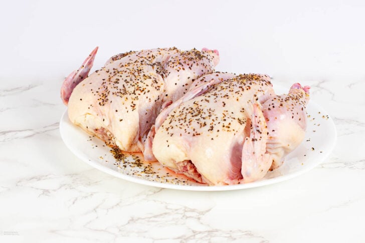 Two raw whole chickens on a white plate, sprinkled with dried herbs, placed on a white marble surface with a plain light background.