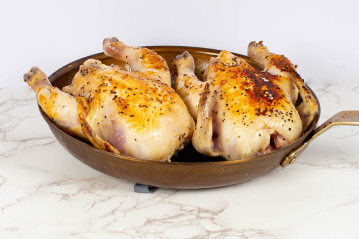 A skillet containing two browned, seasoned spatchcocked chickens sits on a white marble countertop against a plain white background.