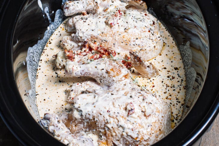 A whole seasoned chicken covered in a creamy sauce with herbs and spices sits inside a slow cooker.