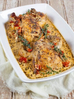 Two roasted cornish hens in a creamy herb sauce, garnished with fresh thyme and sun-dried tomatoes, served in a white rectangular dish on a rustic wooden table.