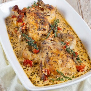 Two roast chickens in a white baking dish with creamy herb sauce, garnished with sprigs of thyme and pieces of sun-dried tomatoes. The dish sits on a light-colored cloth on a rustic wooden table.