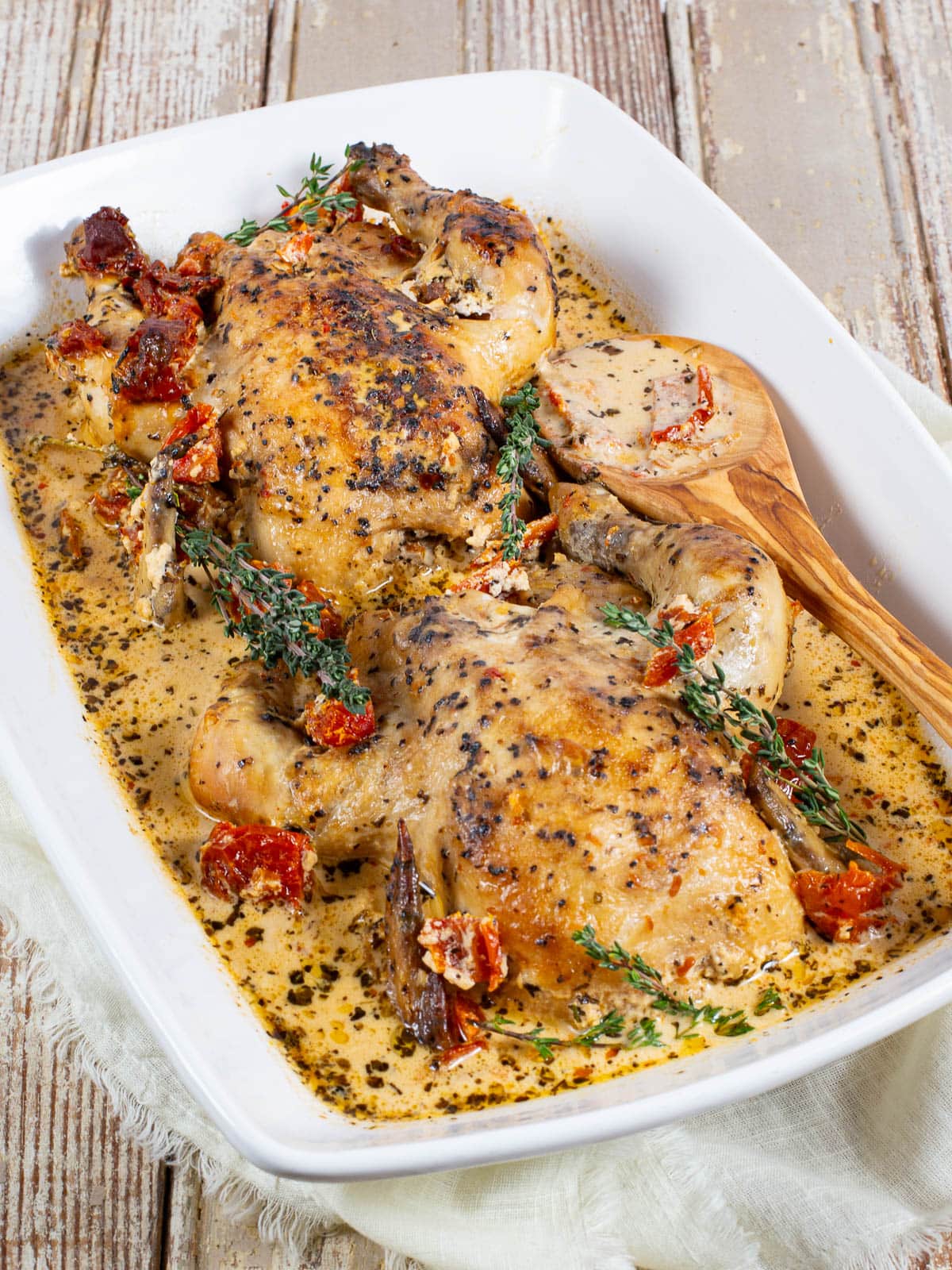 Two cooked chicken breasts in a creamy herb sauce with sun-dried tomatoes, garnished with fresh thyme, served in a white baking dish with a wooden serving spoon on a rustic wooden table.