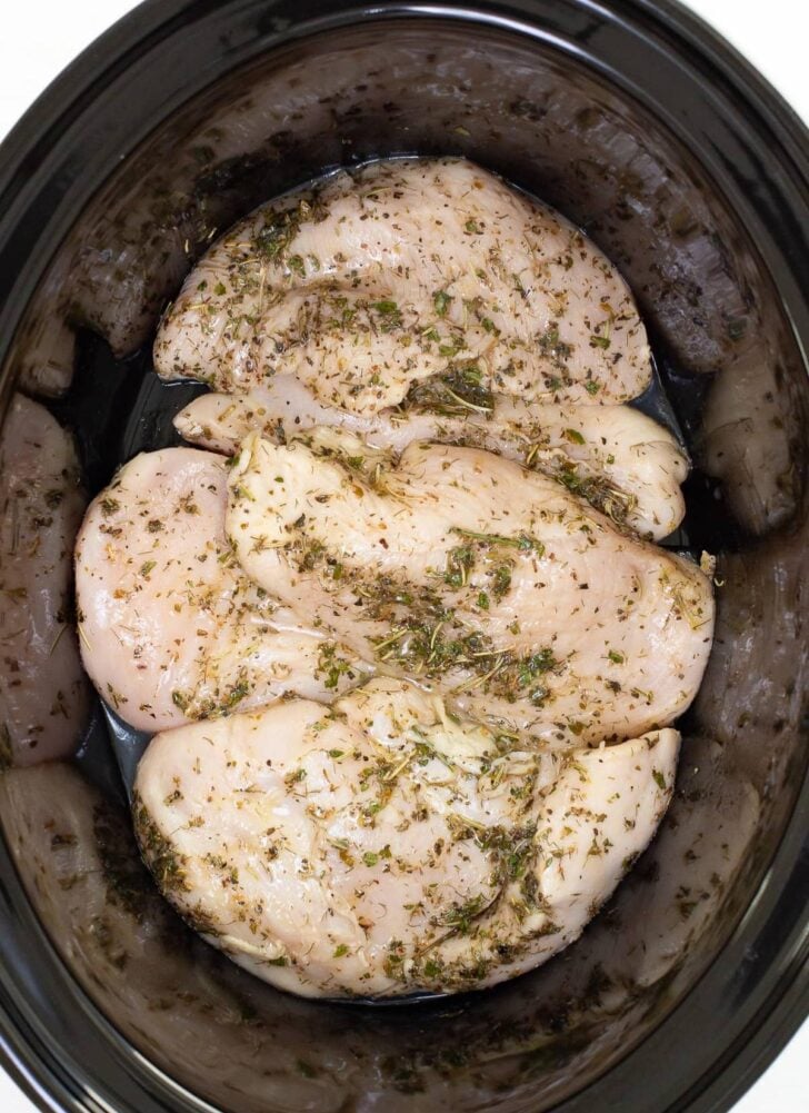 Raw chicken breasts seasoned with herbs and spices are arranged in a slow cooker, ready to be cooked.