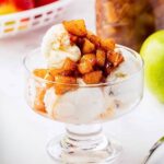 A glass dessert dish filled with scoops of vanilla ice cream, topped with caramelized cinnamon apples. An apple and other food items are blurred in the background.