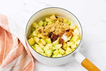 A white saucepan filled with chopped green apples, brown sugar, and spices sits on a white surface next to a pink and white striped kitchen towel.