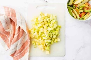Chopped green apples on a white cutting board, next to a bowl of apple peels and cores, and an orange-and-white striped kitchen towel on a white countertop.