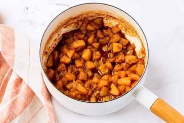 A white saucepan filled with cooked, diced apples in a brown cinnamon sauce sits on a marble surface next to a pink and white striped cloth.