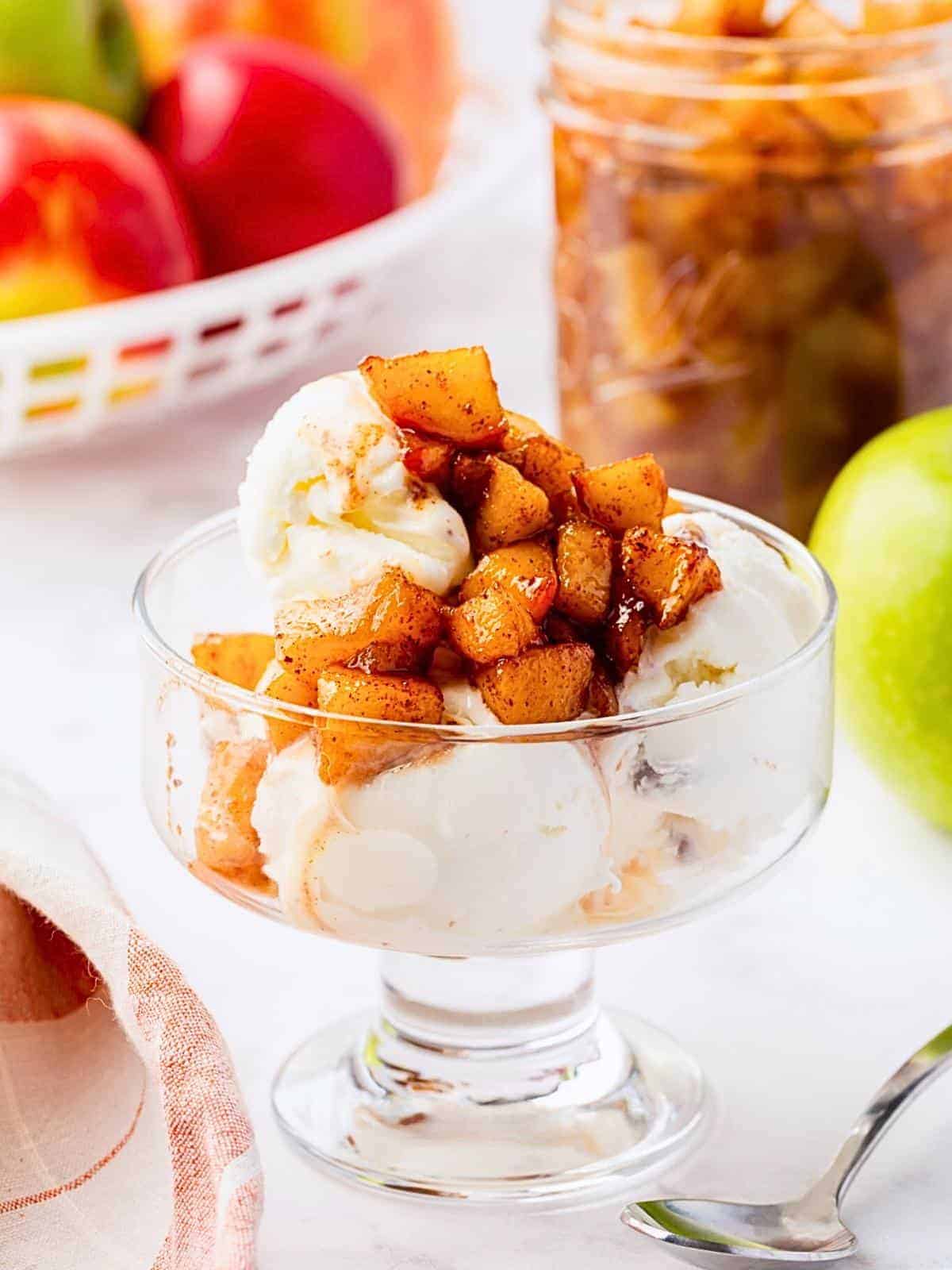 A glass dish of vanilla ice cream topped with caramelized, cinnamon-spiced apple chunks. A jar of apples, a basket of apples, and a green apple are in the background. A spoon and cloth napkin are nearby.