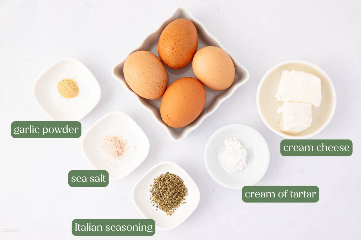 Top-down view of three brown eggs, cream cheese, garlic powder, sea salt, Italian seasoning, and cream of tartar, each in separate white bowls. Labels identify each ingredient.