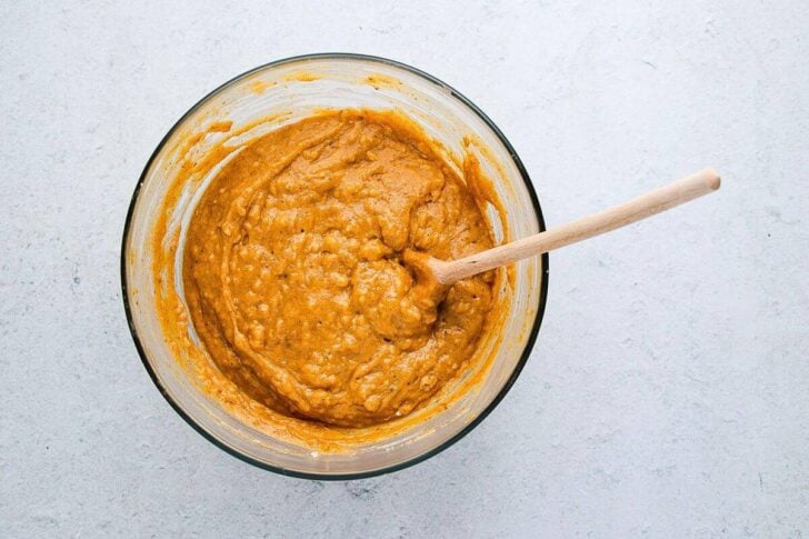 A glass bowl filled with thick, orange-brown batter sits on a light surface. A wooden spoon is resting inside the bowl, partially submerged in the mixture.