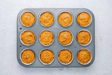 A muffin tin with 12 paper liners, each filled with orange muffin batter, sits on a light-colored surface. The batter appears swirled and is ready for baking.
