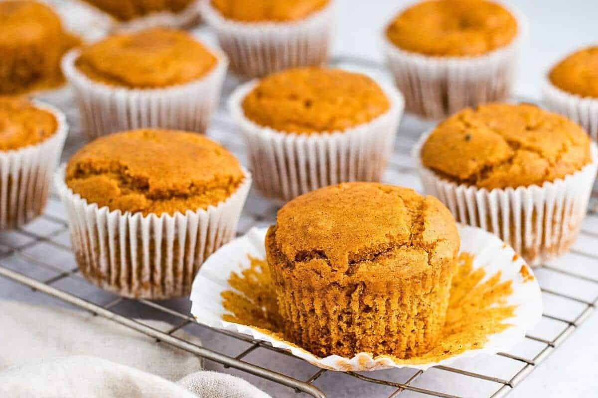 A batch of golden-brown muffins in white paper liners is displayed on a wire cooling rack. One muffin in the foreground has its liner partially peeled back, revealing its soft texture.