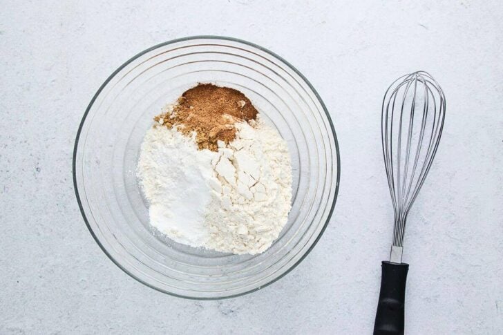 A glass bowl with flour, baking powder, and cinnamon sits on a light surface next to a metal whisk with a black handle.