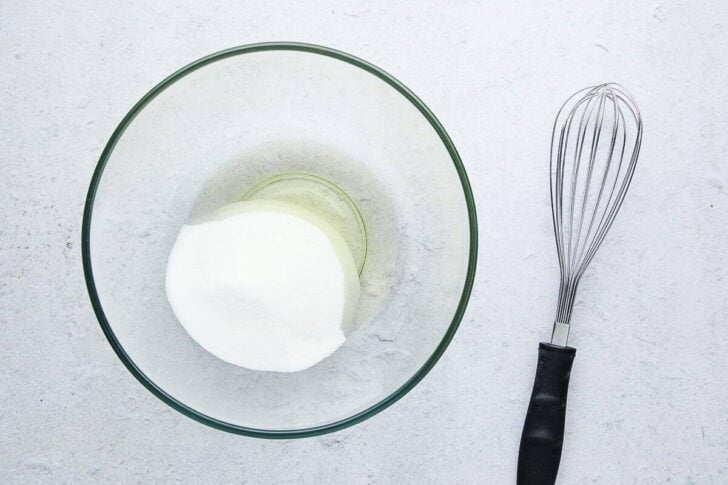 A glass bowl with egg whites and sugar inside, placed on a light surface next to a metal whisk with a black handle.