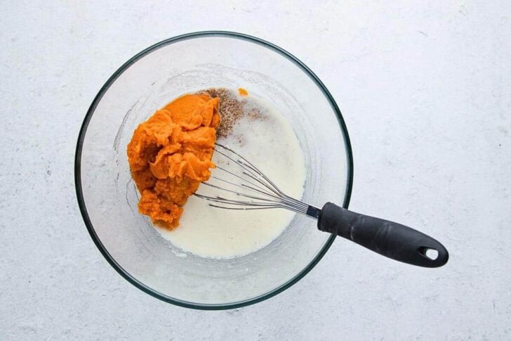 A glass bowl containing pumpkin puree, milk, and spices with a whisk resting inside, placed on a white surface.