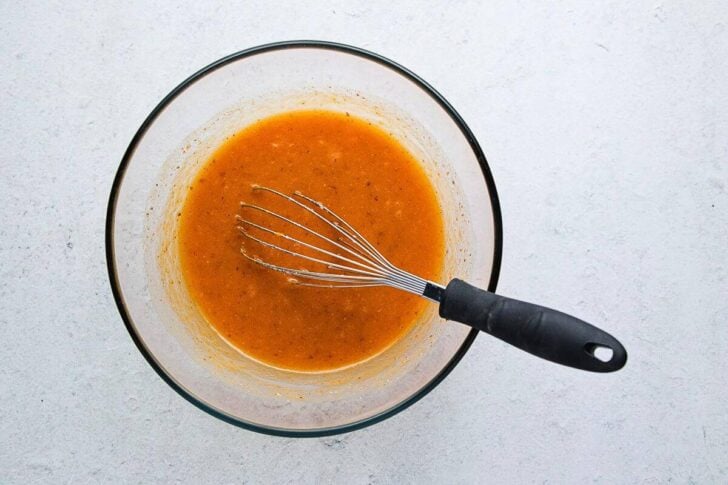 A glass bowl filled with an orange-brown liquid, possibly a sauce or dressing, with a metal whisk resting inside on a light gray surface.